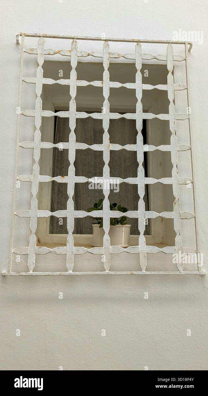 Minimalistisches weißes Fenster mit dekorativem geometrischem Eisengitter und kleiner Topfpflanze an weiß getünchter Wand - Smartphone-aufgenommenes Stockfoto