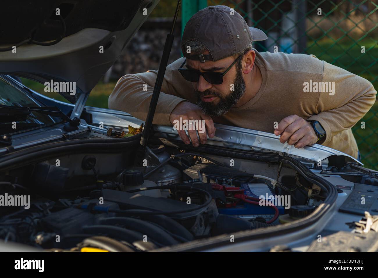 Ein Mann beugt sich über die geöffnete Motorhaube eines Autos und untersucht die Motorteile, während er eine Sonnenbrille trägt. Es ist tagsüber und die Umgebung ist draußen. Stockfoto