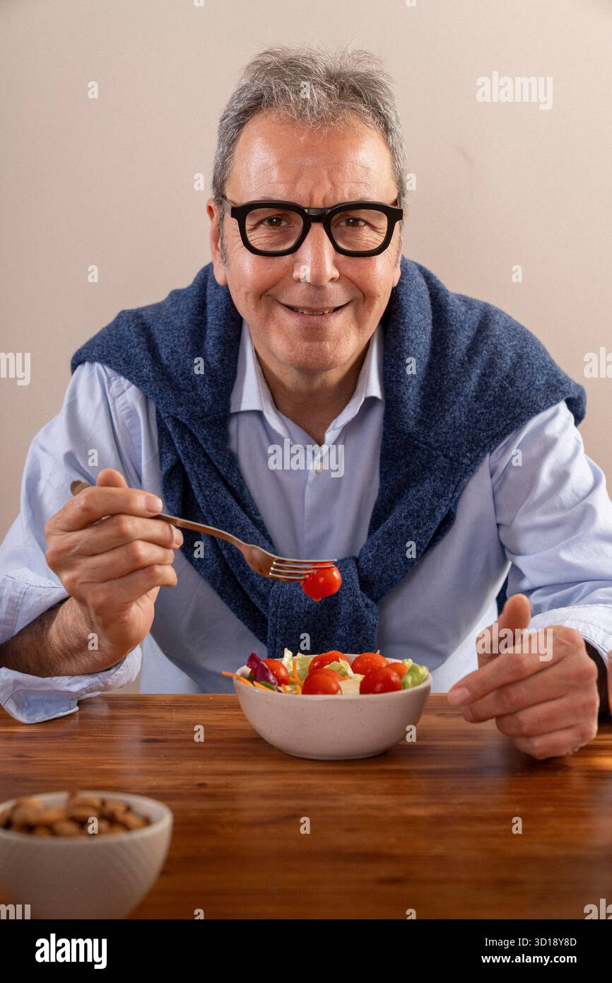 Ein fröhlicher Mann sitzt an einem Holztisch und genießt einen bunten Salat. Er trägt einen blauen Pullover über den Schultern und lächelt warm und sorgt für eine Freude Stockfoto