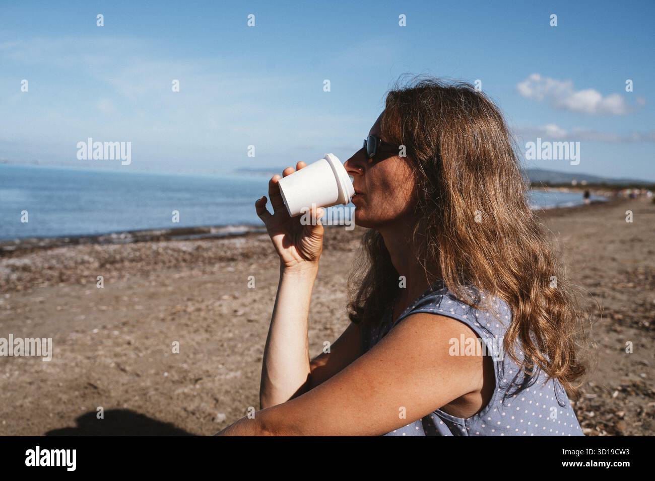 Eine Frau schlürft an einem Becher, während sie an einem Sandstrand sitzt. Der ruhige Blick auf das Meer schafft eine ruhige Umgebung, perfekt für Entspannung und introspektive Momente Stockfoto