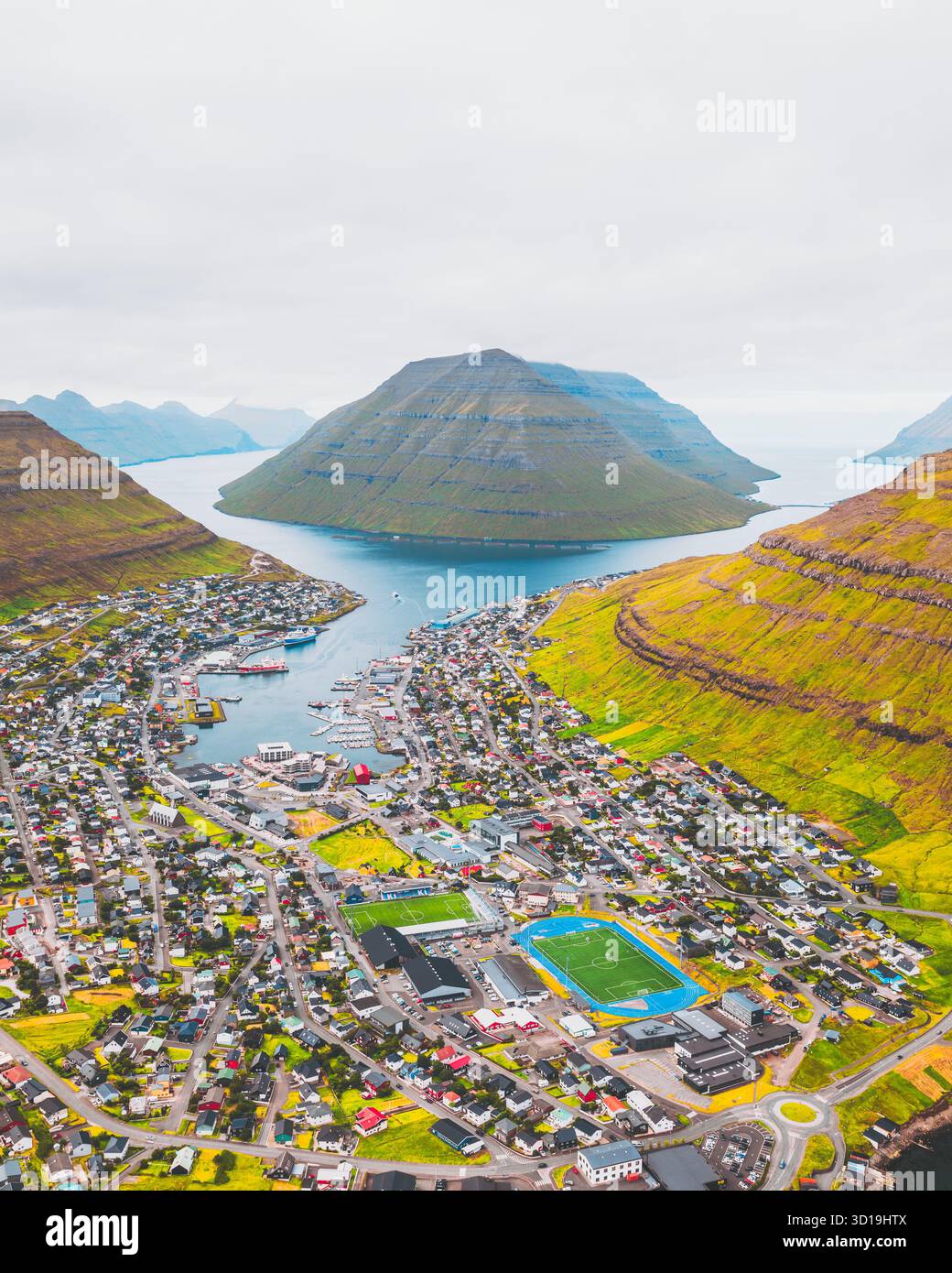 Blick aus der Vogelperspektive auf Klaksvík, eingebettet zwischen majestätischen grünen Hügeln und dem blauen Meer, mit der weitläufigen Stadt und dem Fußballfeld, die lebendige Spritzer von c Stockfoto