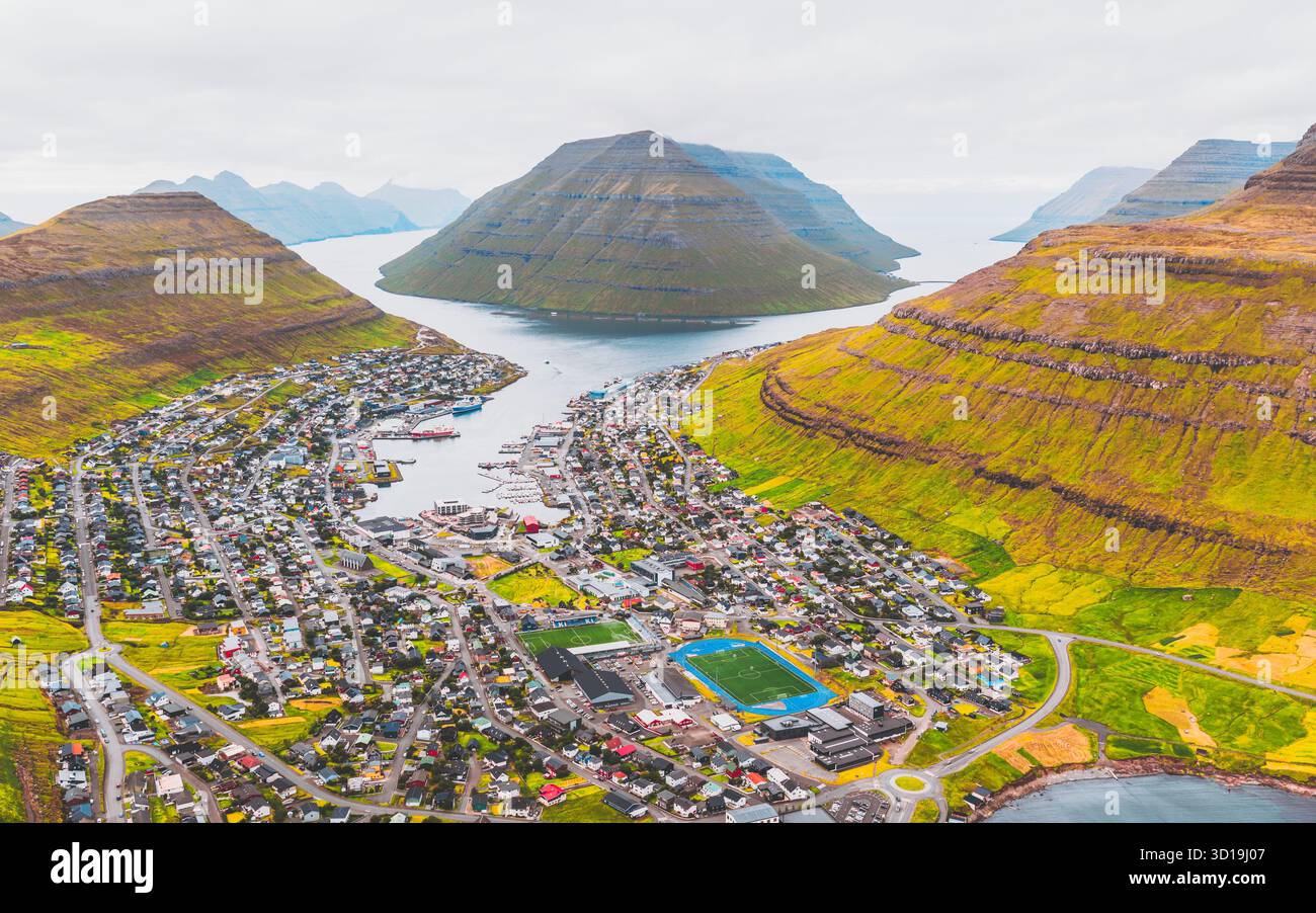 Aus der Vogelperspektive von Klaksvík eingebettet zwischen dramatischen grünen Hügeln und dem klaren blauen Meer, eine lebendige Stadt unter einem weichen, bedeckten Himmel, Klaksvík, Nördliche Inseln, Färöer Inseln. Stockfoto