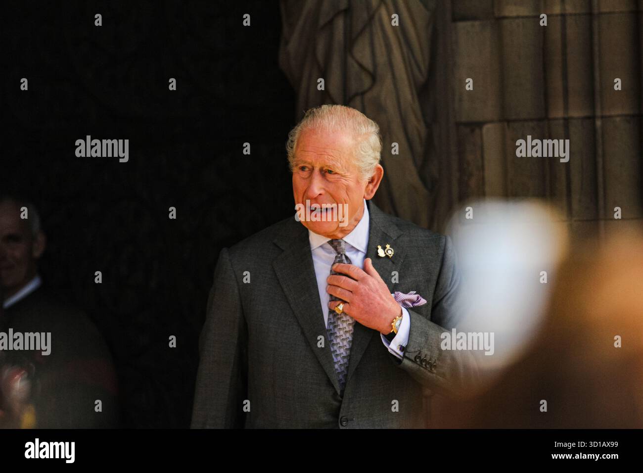 König Karl III. Passt seine Verbindung während eines öffentlichen Auftritts in Lichfield, Staffordshire, an. König Karl III. Wird in einem formellen dunkelgrauen Anzug mit einem lavendelfarbenen Taschenquadrat und gemusterter Krawatte vor einer historischen Steinmauer in Lichfield gesehen. Der Monarch scheint sich auf eine zeremonielle Verlobung vorzubereiten, während andere Teilnehmer im Hintergrund sichtbar sind. Die Umgebung empfiehlt einen offiziellen Besuch eines Kulturerbes oder eines öffentlichen Ortes. Stockfoto