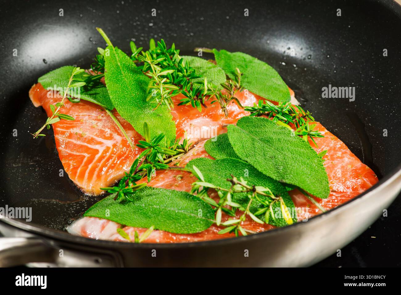 Erhöhte Sicht auf zwei Lachsfilets, die mit frischem Salbei und Thymian in einer schwarz beschichteten Pfanne mariniert sind. Stockfoto