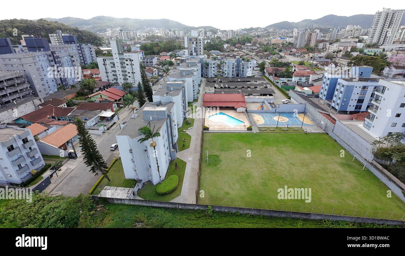 Aus der Vogelperspektive auf eine Wohngegend mit vielen Gebäuden, einem großen Swimmingpool, Fußball- und Futsal-Feldern, umgeben von Bäumen und Bergen unter einem Blau Stockfoto