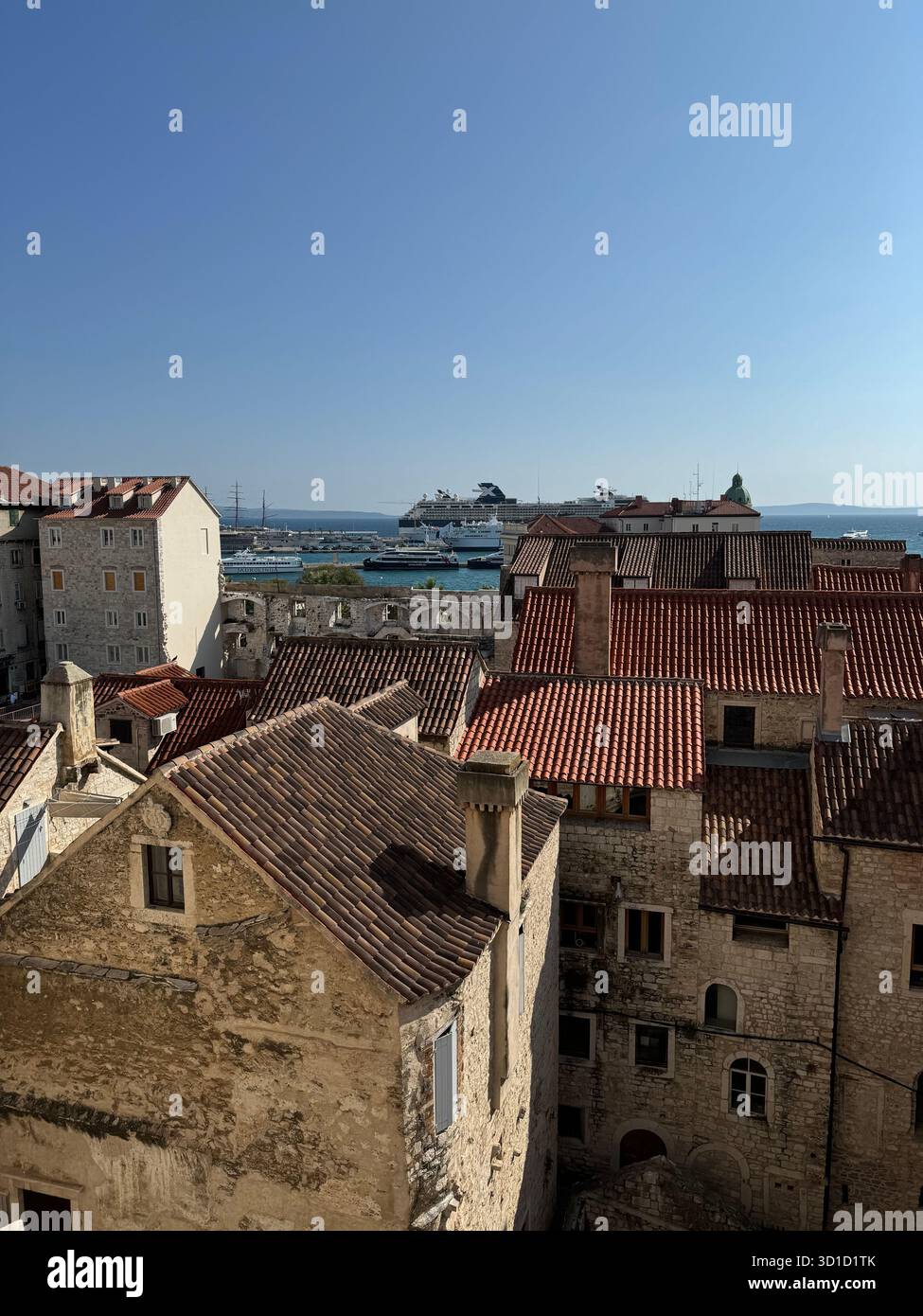 Aus der Vogelperspektive über historische Steinhäuser mit Terrakottafliesen mit Blick auf einen geschäftigen Mittelmeerhafen mit Kreuzfahrtschiffen und die Adria - Smartphone-aufgenommenes Stockfoto