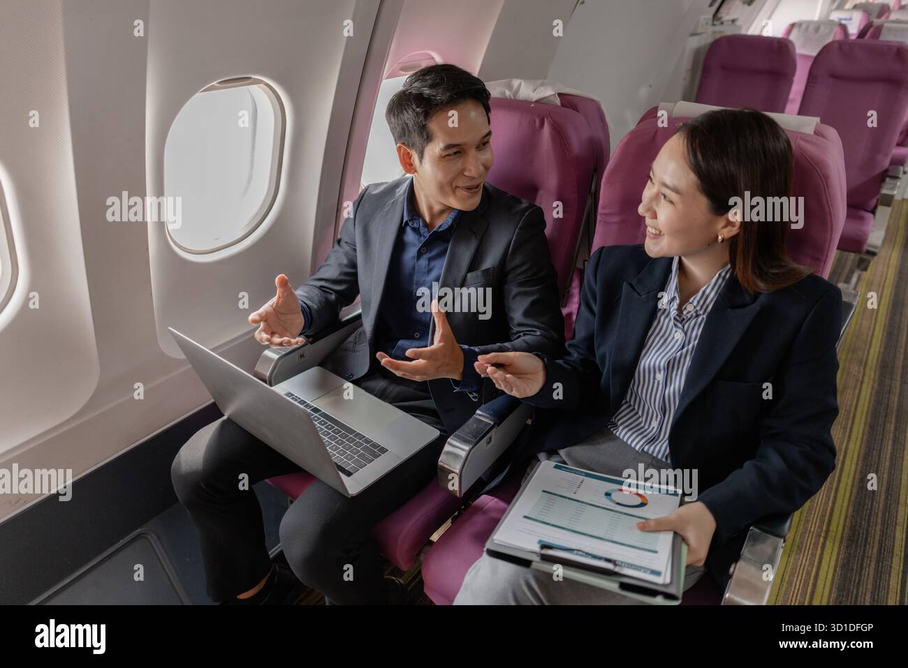 Ein Geschäftsmann und eine Frau in formellen Anzügen arbeiten zusammen auf einem Flug. Sie diskutieren Dokumente und verwenden Laptops, um Teamarbeit und Produktivität zu zeigen Stockfoto