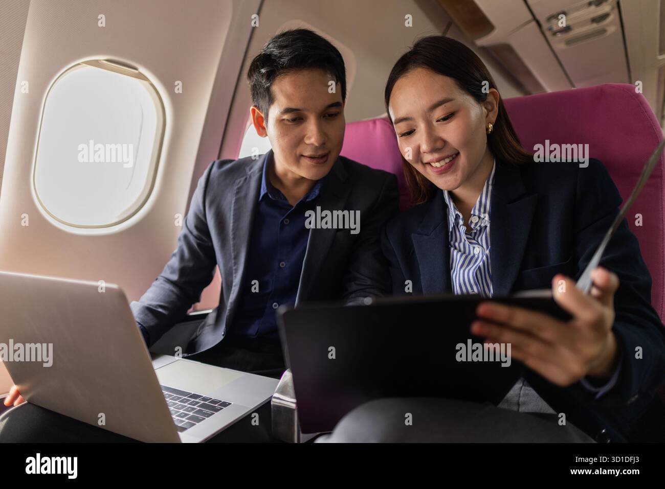 Ein Geschäftsmann und eine Frau in formellen Anzügen arbeiten zusammen auf einem Flug. Sie diskutieren Dokumente und verwenden Laptops, um Teamarbeit und Produktivität zu zeigen Stockfoto