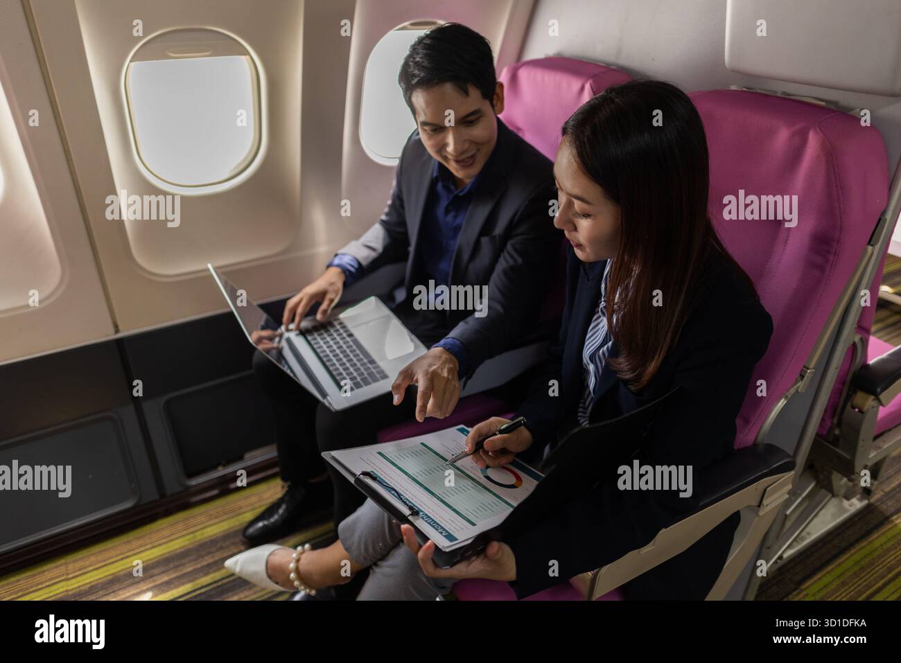 Ein Geschäftsmann und eine Frau in formellen Anzügen arbeiten zusammen auf einem Flug. Sie diskutieren Dokumente und verwenden Laptops, um Teamarbeit und Produktivität zu zeigen Stockfoto