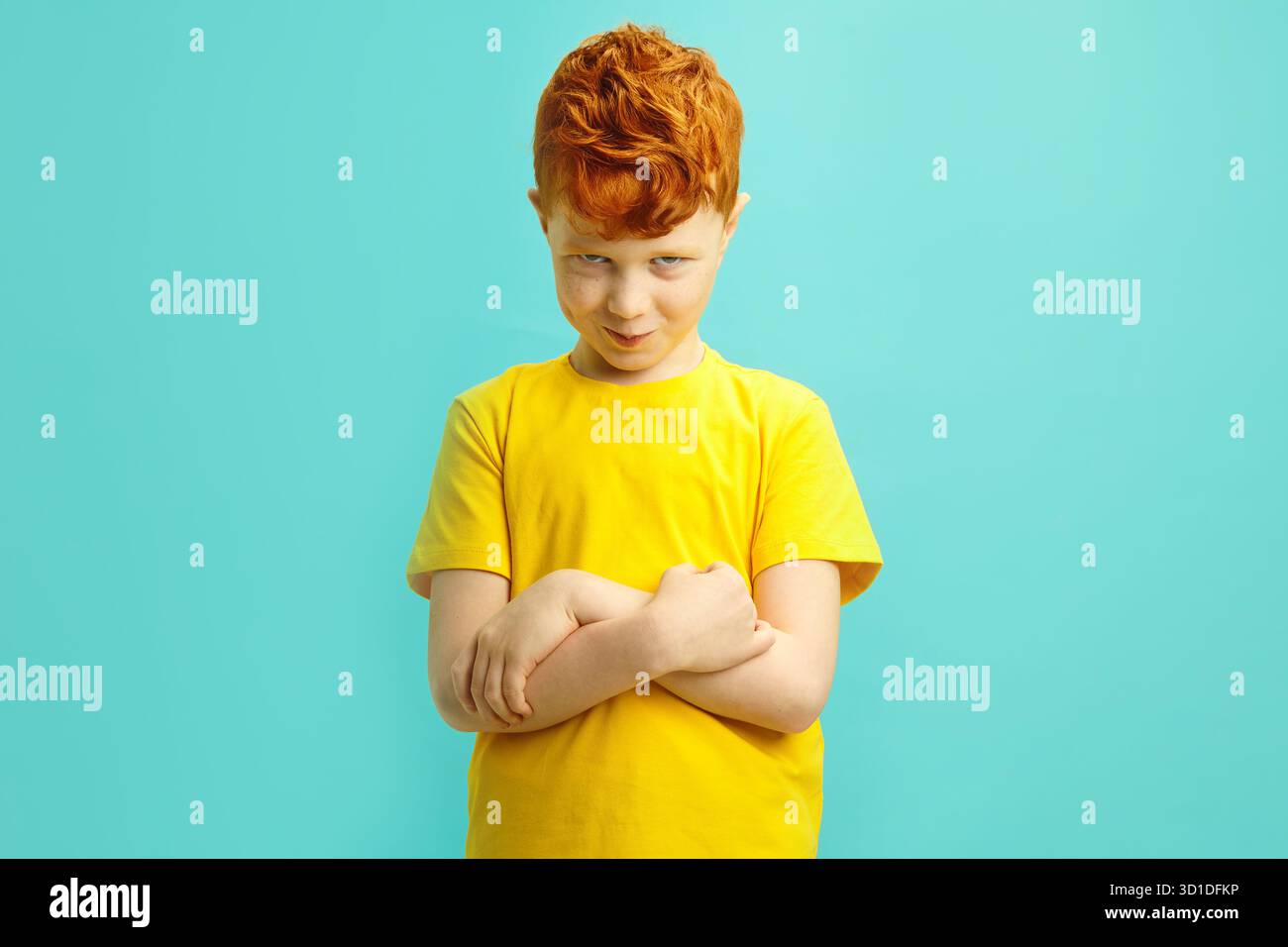 Der freche rothaarige Junge etwa 7-8 Jahre kreuzt trotzig die Arme vor ihm, nachdem er das Gesicht beleidigt hat, das auf blauem isoliertem Hintergrund steht. Stockfoto