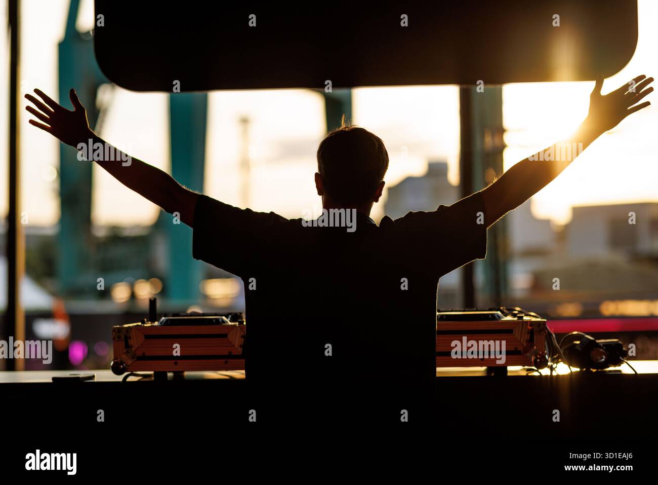 Silhouette eines DJ, der hinter der Ausrüstung steht und die Arme gegen helles Sonnenuntergangslicht am Musiklokal im Freien angehoben sind. Stockfoto