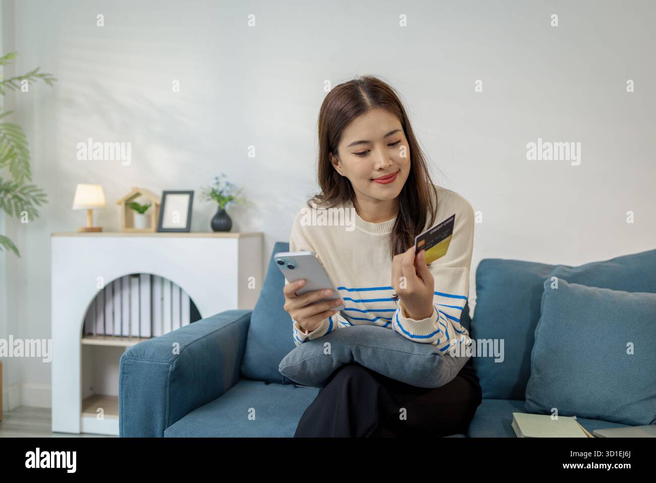 Eine junge Frau sitzt auf einem Sofa und hält ein Smartphone und eine Kreditkarte und lächelt, während sie eine Online-Transaktion tätigt. Sie sieht entspannt und selbstbewusst aus Stockfoto
