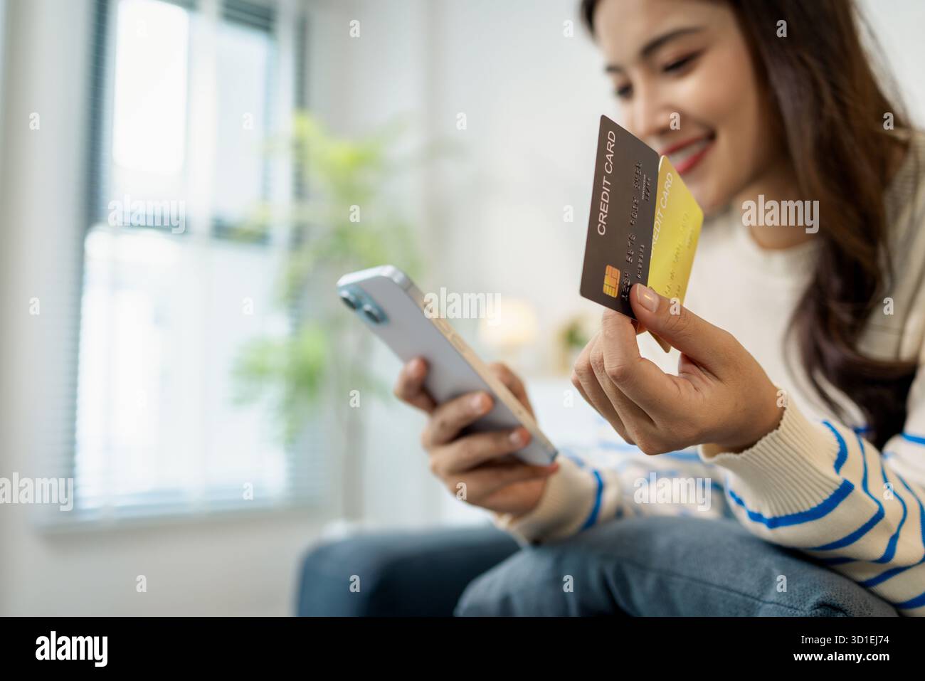 Eine junge Frau sitzt auf einem Sofa und hält ein Smartphone und eine Kreditkarte und lächelt, während sie eine Online-Transaktion tätigt. Sie sieht entspannt und selbstbewusst aus Stockfoto