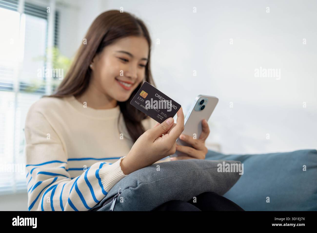 Eine junge Frau sitzt auf einem Sofa und hält ein Smartphone und eine Kreditkarte und lächelt, während sie eine Online-Transaktion tätigt. Sie sieht entspannt und selbstbewusst aus Stockfoto