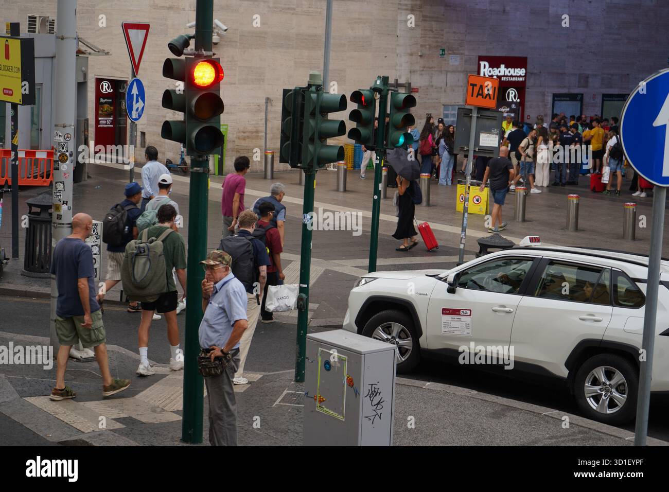 Ein belebter Taxistand in Rom, mit einem weißen SUV-Taxi neben Fußgängern und verschiedenen Ampeln, was auf ein geschäftiges Stadtviertel hinweist. Stockfoto