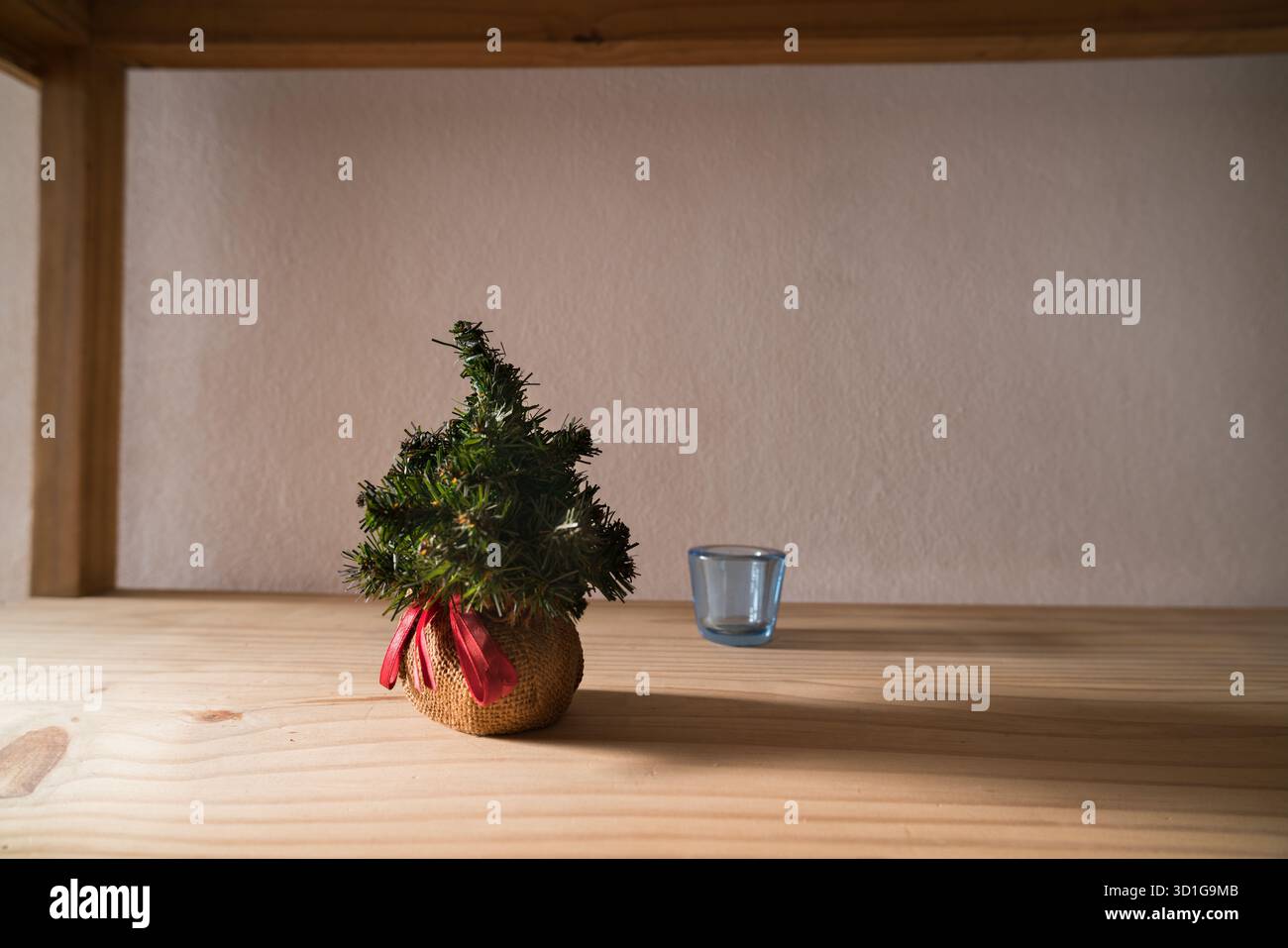 Auf einem hellen Holzregal steht ein bezaubernder Miniatur-Weihnachtsbaum, eingebettet in einen Sackleinen mit rotem Band. Daneben ein kleines leeres Glas Stockfoto