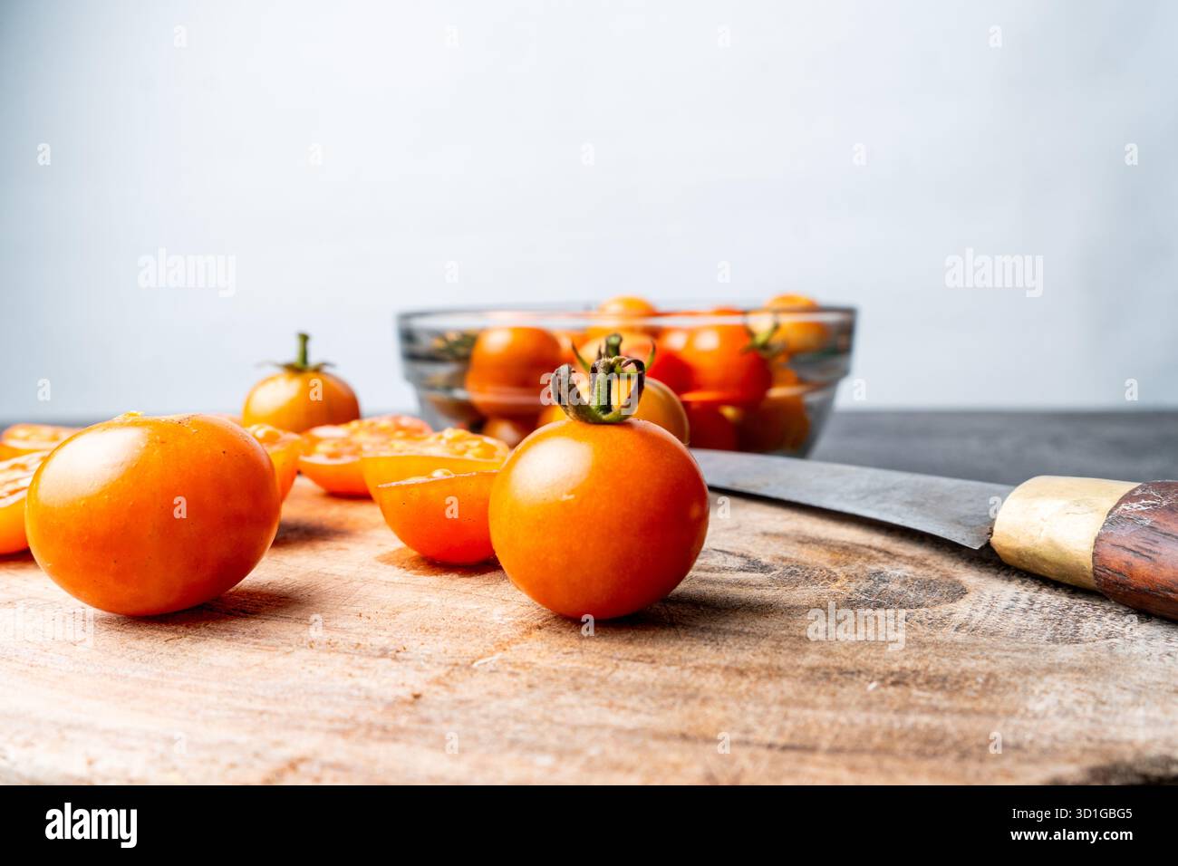 Nahaufnahme von frischen Kirschtomaten, ganz und in Scheiben geschnitten, auf einem rustikalen Holzschneidebrett. Ein Messer, eine Schüssel und Limetten sind im weichen Bokeh-Hintergrund. Stockfoto