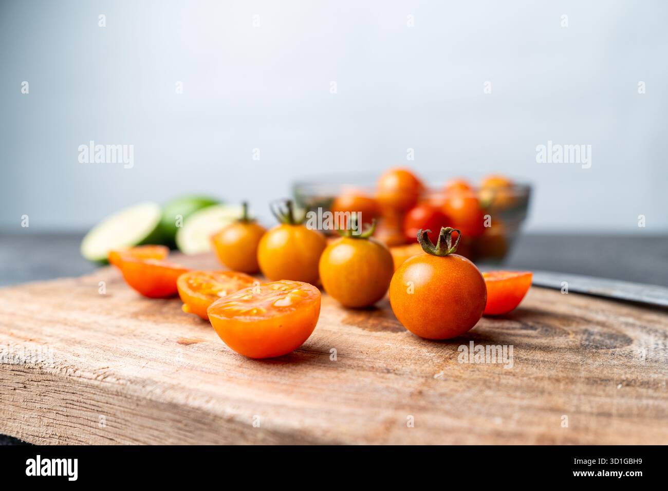 Nahaufnahme von frischen Kirschtomaten, ganz und in Scheiben geschnitten, auf einem rustikalen Holzschneidebrett. Ein Messer, eine Schüssel und Limetten sind im weichen Bokeh-Hintergrund. Stockfoto