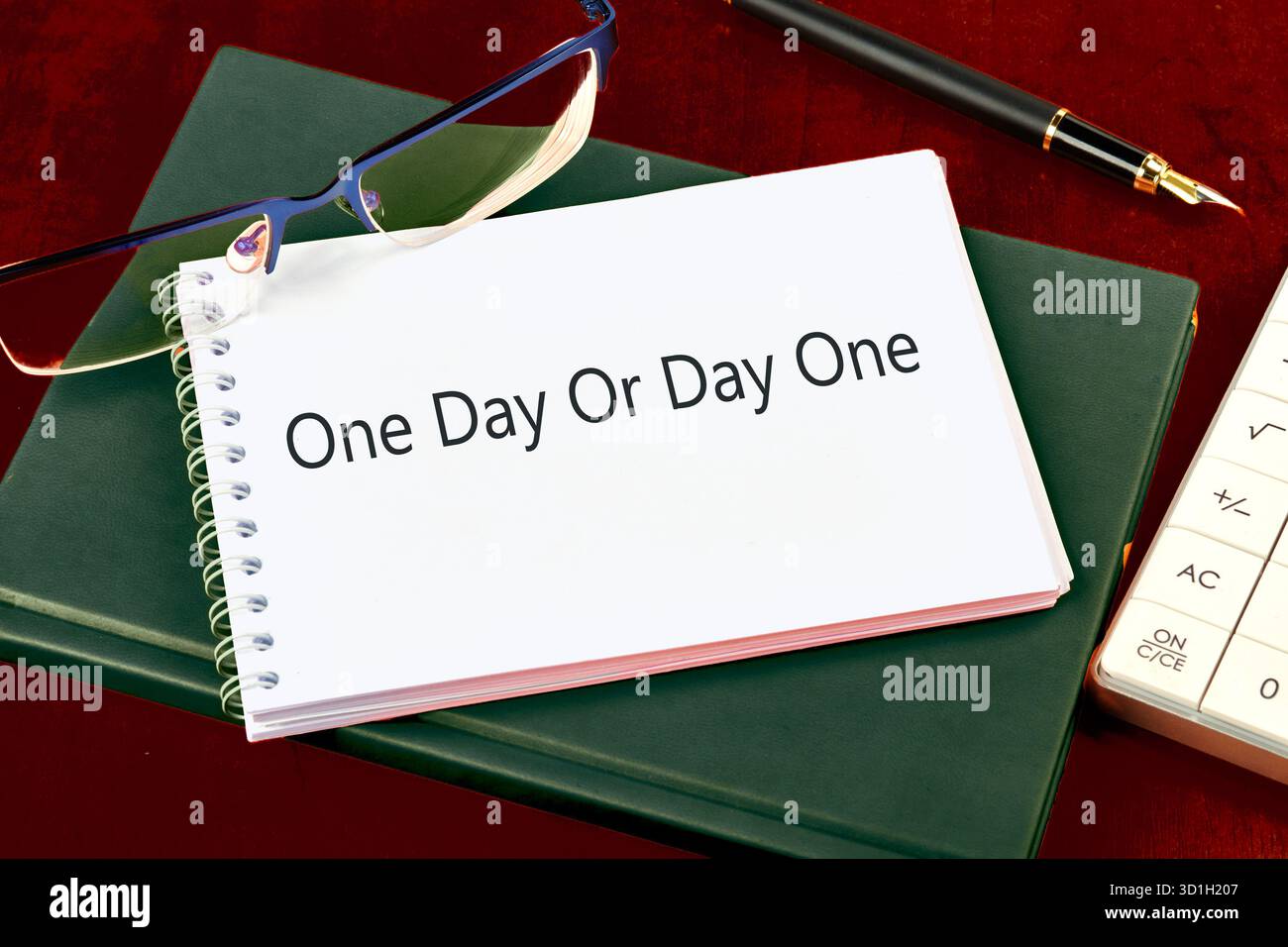 Ein Notizbuch mit dem Satz One Day oder Day One liegt auf einem grünen Buch. Daneben wartet eine Brille auf Sie, die einen Moment tiefer Gedanken und Reflektionen signalisiert Stockfoto