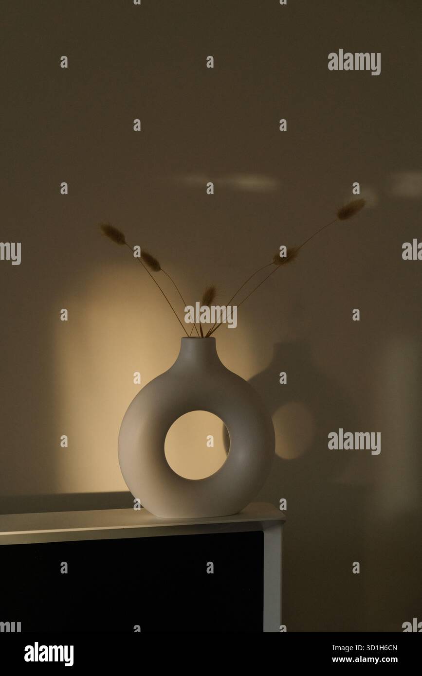 Minimale runde Keramikvase mit getrockneten Stielen in warmem Sonnenuntergangslicht und weichen Wandschatten, ruhiges und elegantes Stillleben im Innenraum. Stockfoto