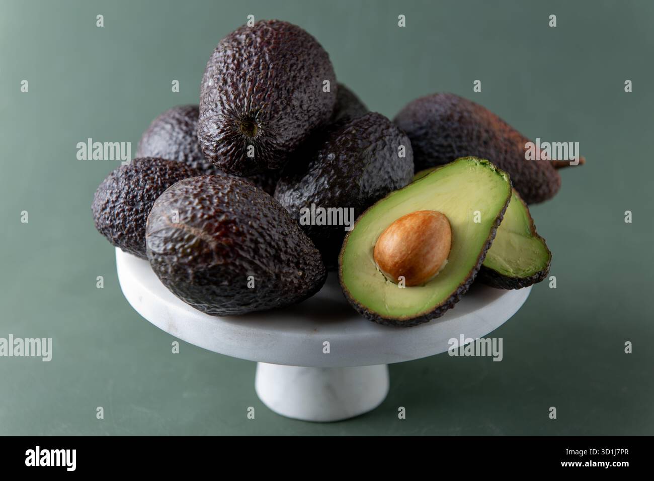 Reife Hass Avocados auf einem weißen Marmorständer, wobei einer offen geschnitten ist, um das grüne Fleisch und den Samen vor einem weichen grünen Hintergrund zu zeigen. Stockfoto