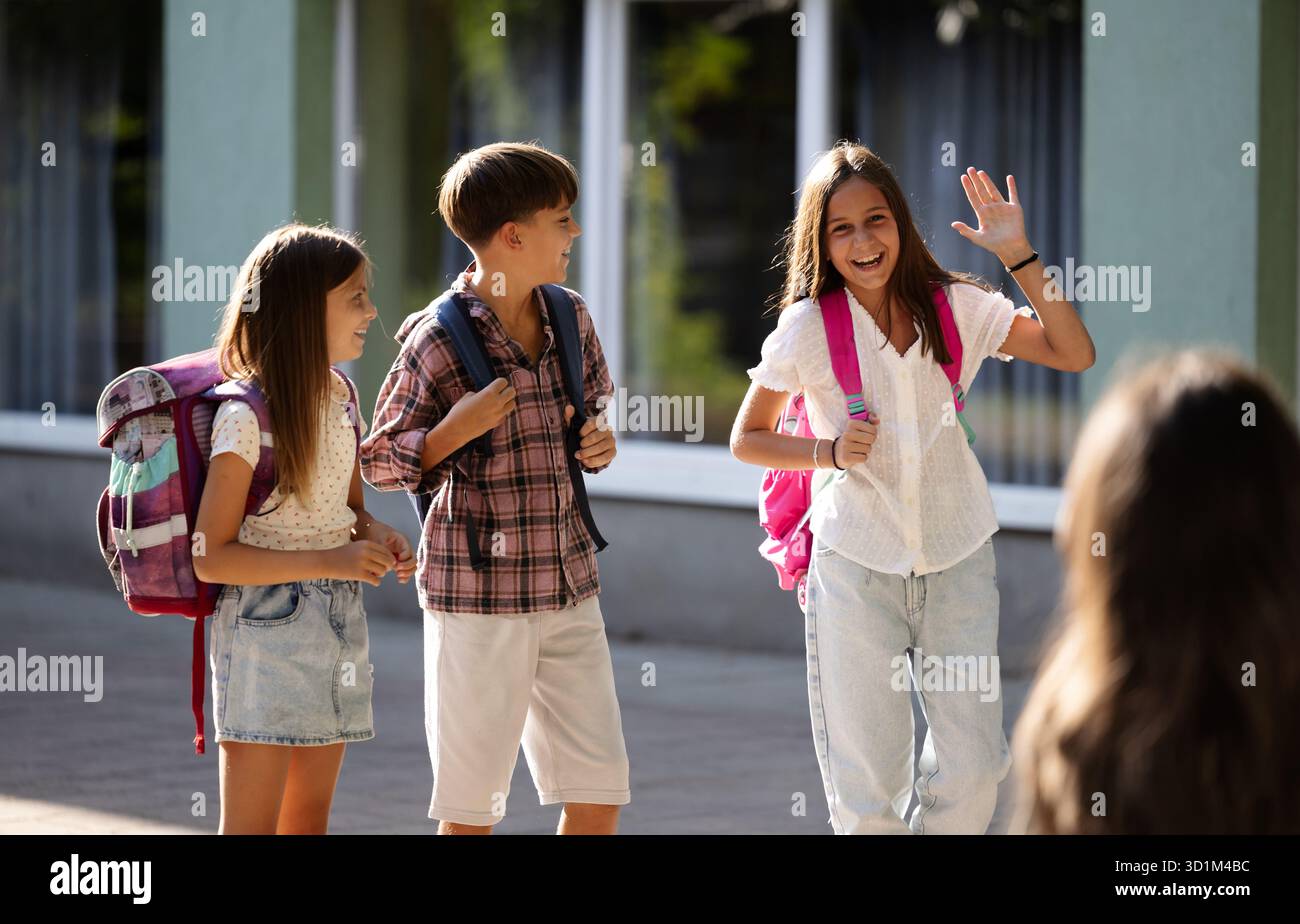 Ein fröhliches Mädchen winkte mit der Hand in der Begrüßung, als sie ihre Mutter nach dem Unterricht sah und mit ihren Freunden aus der Schule ging Stockfoto