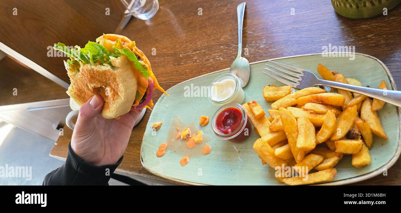 Hand hält einen frischen Burger mit Salat und Belag, begleitet von knusprigen Pommes und Saucen auf einem rustikalen Holztisch. - Smartphone-aufgenommenes Stockfoto