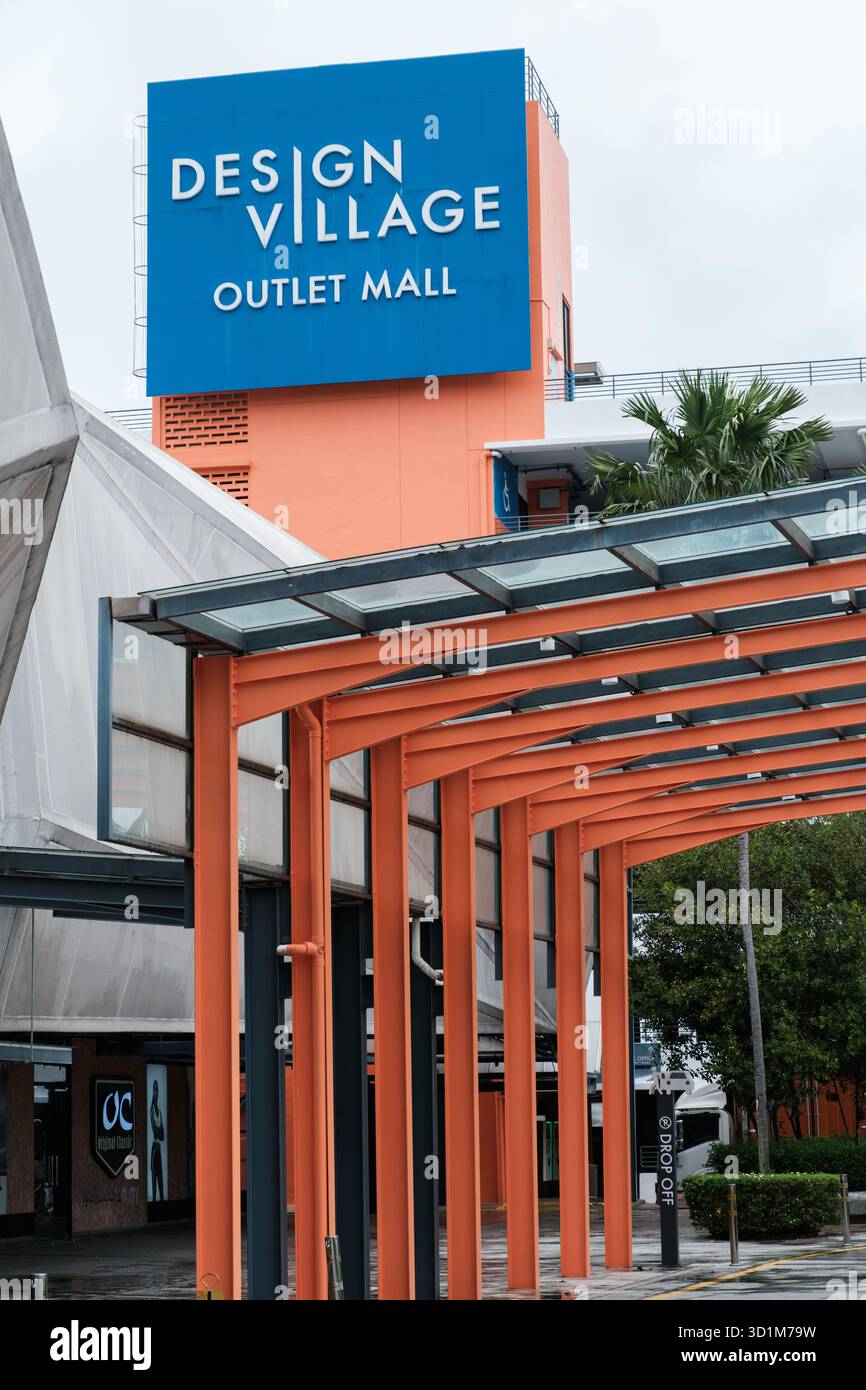 Das Design Village Outlet Mall ist mit einem hellblauen Schild vor einem modernen, orangen Gebäude umgeben von üppigem Grün sichtbar. Stockfoto