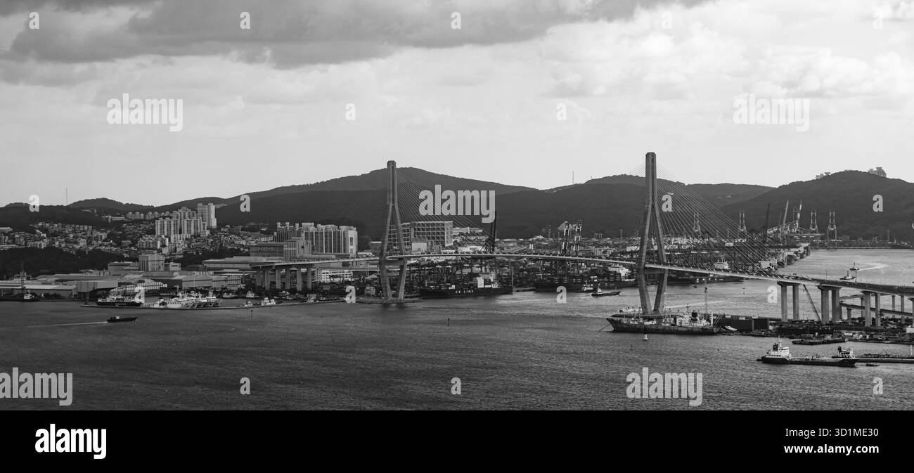 BUSAN, SÜDKOREA - 9. OKTOBER 2025 - Busan Hafen Stadtlandschaft mit Hängebrücke, Frachtschiffen und Containerkränen in Schwarz-weiß Stockfoto