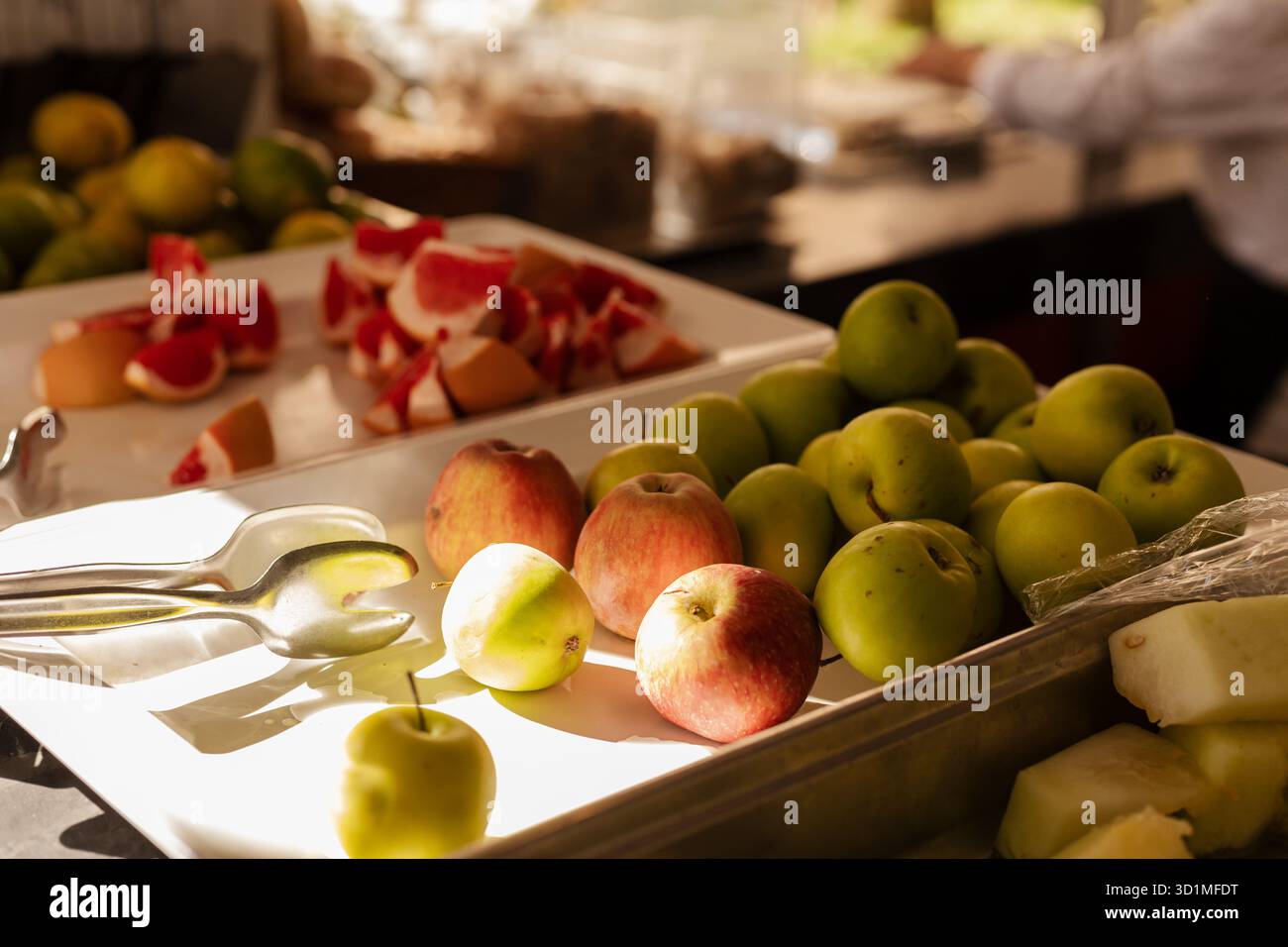 Eine Auswahl an frischem Obst, darunter Äpfel und Grapefruit, werden kunstvoll auf einem Buffettisch arrangiert. Die Umgebung ist hell mit Morgensonne, verbessert Stockfoto