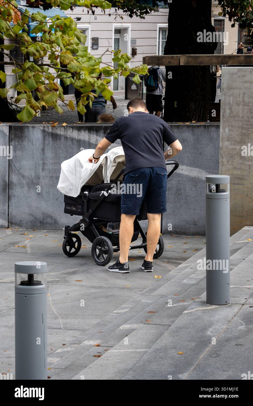 Ljubljana, Slowenien 30. August 2025 Mann, der den Kinderwagen auf Betontreppen schiebt, Bäume im Hintergrund Stockfoto