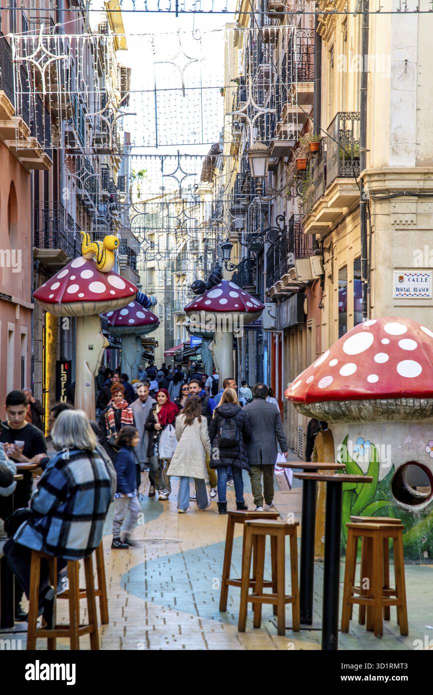 Alicante, Alicante - Spanien - 01-07-2024: Die Gasse von Alicante ist lebhaft mit Fußgängern, skurrilen Kunstinstallationen und Hängeleuchten Stockfoto