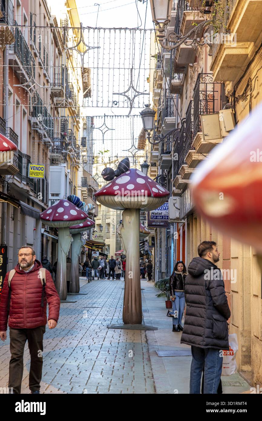 Alicante, Alicante - Spanien - 01-07-2024: Die Gasse von Alicante ist lebhaft mit Fußgängern, skurrilen Kunstinstallationen und Hängeleuchten Stockfoto
