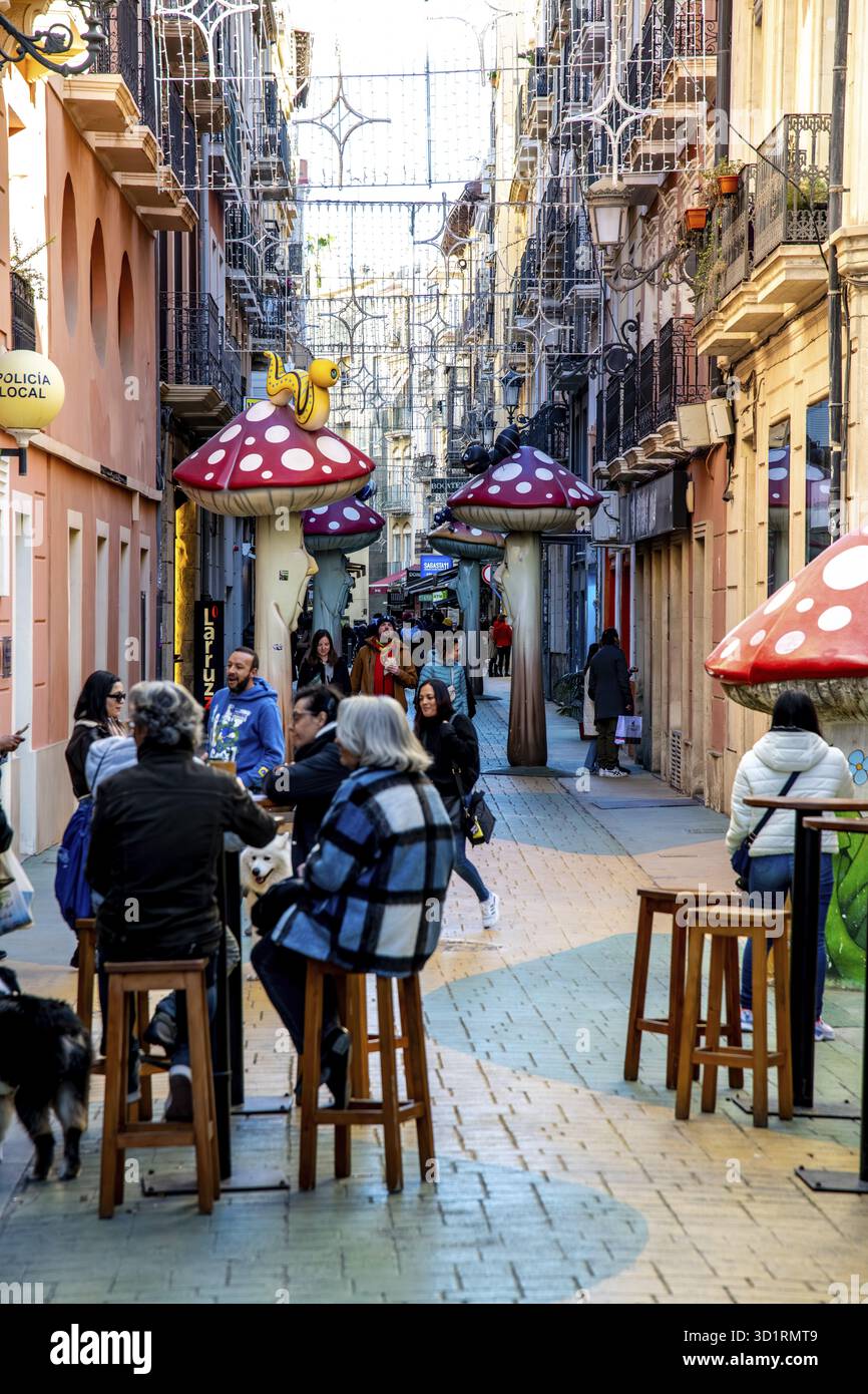 Alicante, Alicante - Spanien - 01-07-2024: Die Gasse von Alicante ist lebhaft mit Fußgängern, skurrilen Kunstinstallationen und Hängeleuchten Stockfoto