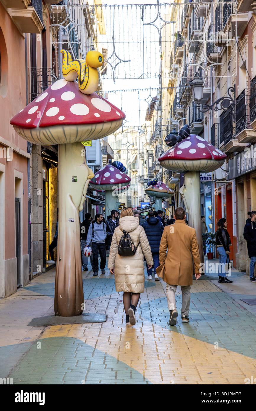 Alicante, Alicante - Spanien - 01-07-2024: Die Gasse von Alicante ist lebhaft mit Fußgängern, skurrilen Kunstinstallationen und Hängeleuchten Stockfoto