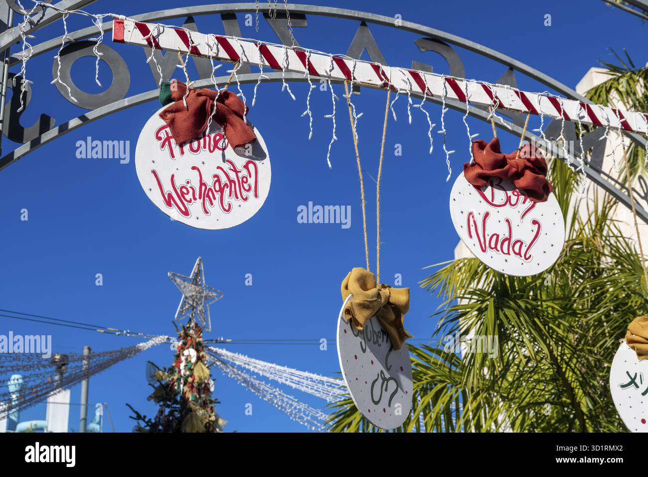 Denia, Alicante - Spanien - 22.12.2023: Hellblauer Himmel mit „Merry Christmas“-Schild in verschiedenen Sprachen und rotem Bogen Stockfoto
