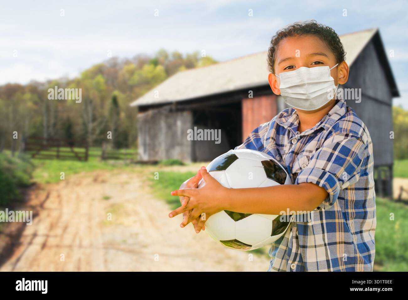 Mixed Race Hispanic und Afro American Boy halten Fußball und tragen Gesichtsmaske im Freien Stockfoto