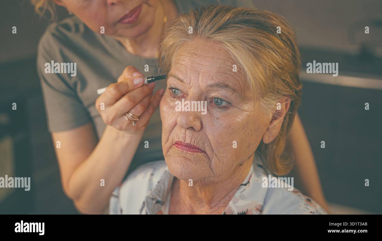 Zeitlose Eleganz: Eine junge Kosmetikerin veredelt eine über 85+ blonde Frau mit blauen Augen mit Mascara, um ihre Wimpern zu betonen Stockfoto