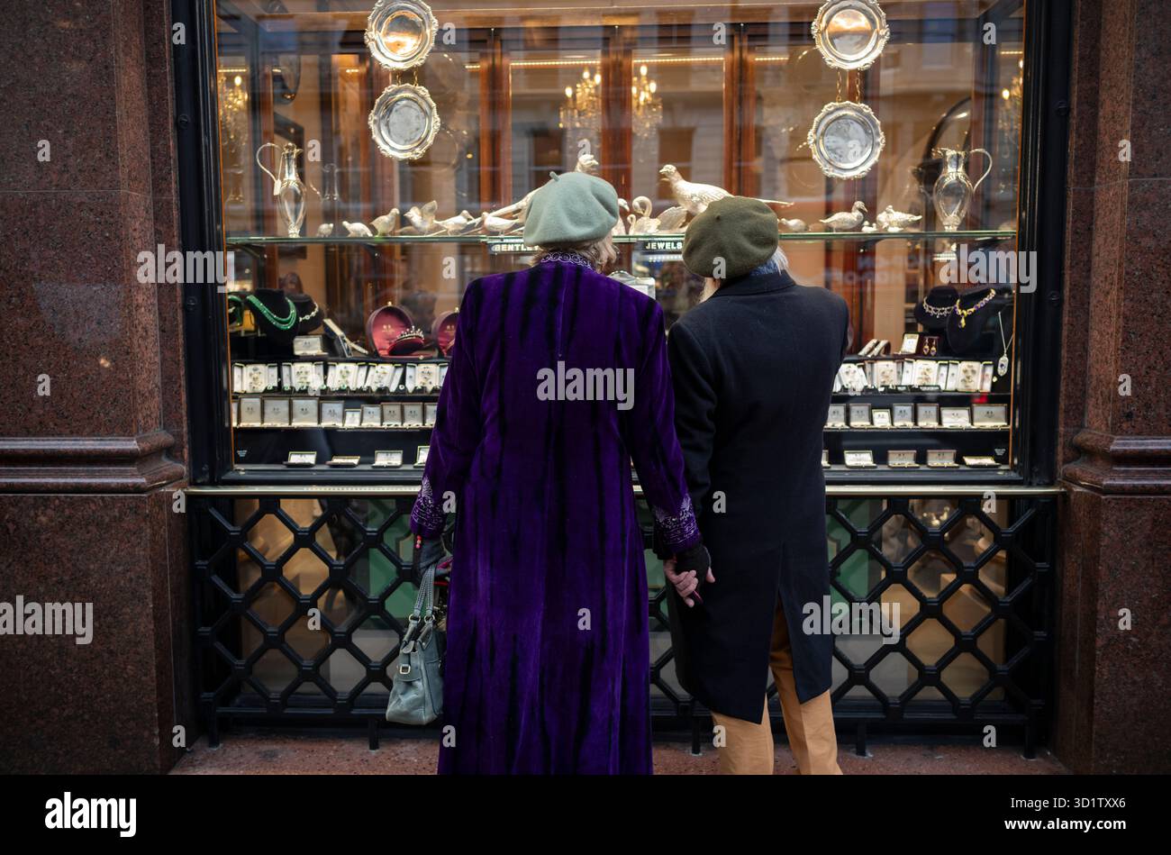 Ein Paar mit Baskenmützen und Wintermänteln bietet einen Blick auf Bentley & Skinner, Mayfair Antiquitätenausstellung und maßgeschneiderte Schmuckgeschäfte in Piccadilly, London, Großbritannien. Stockfoto