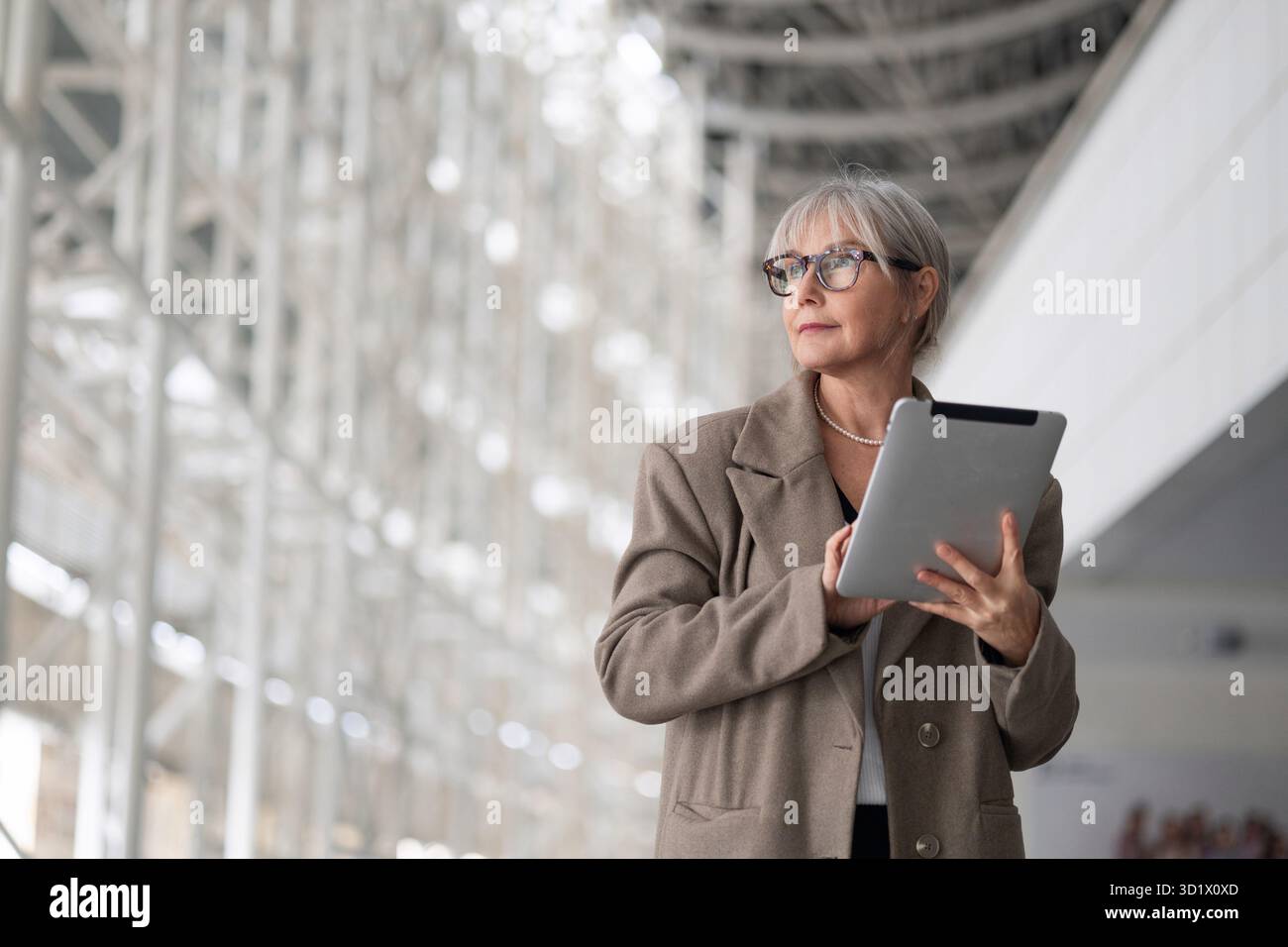 Stilvolle ältere Frau mit Brille, die ein Tablet in einem modernen, gut beleuchteten architektonischen Raum am Nachmittag verwendet Stockfoto