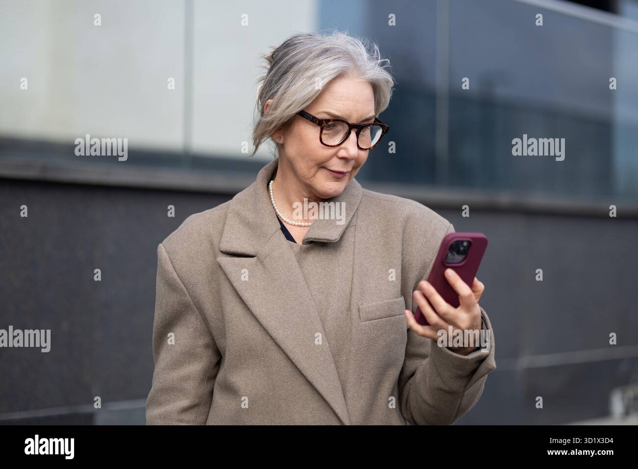 Eine ältere Geschäftsfrau überprüft ihr Smartphone, während sie an einem sonnigen Nachmittag vor einem modernen Bürogebäude steht Stockfoto