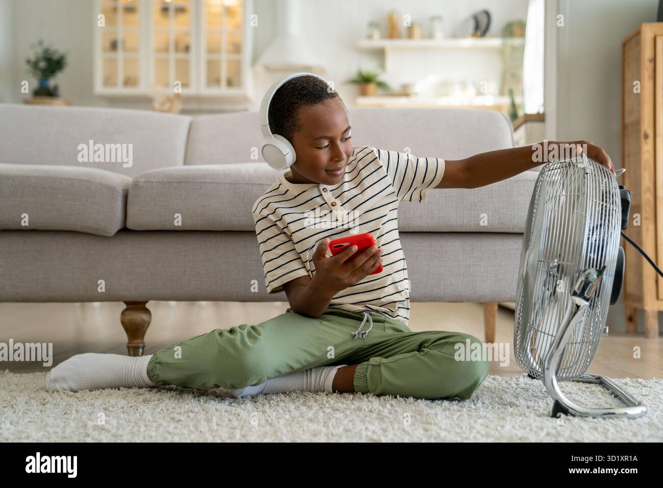 Ein zufriedener gemischter Junge, der auf dem Teppich sitzt und mit einem elektrischen Ventilator auf dem Boden ausgestattet ist, hört Musik auf dem Handy Stockfoto