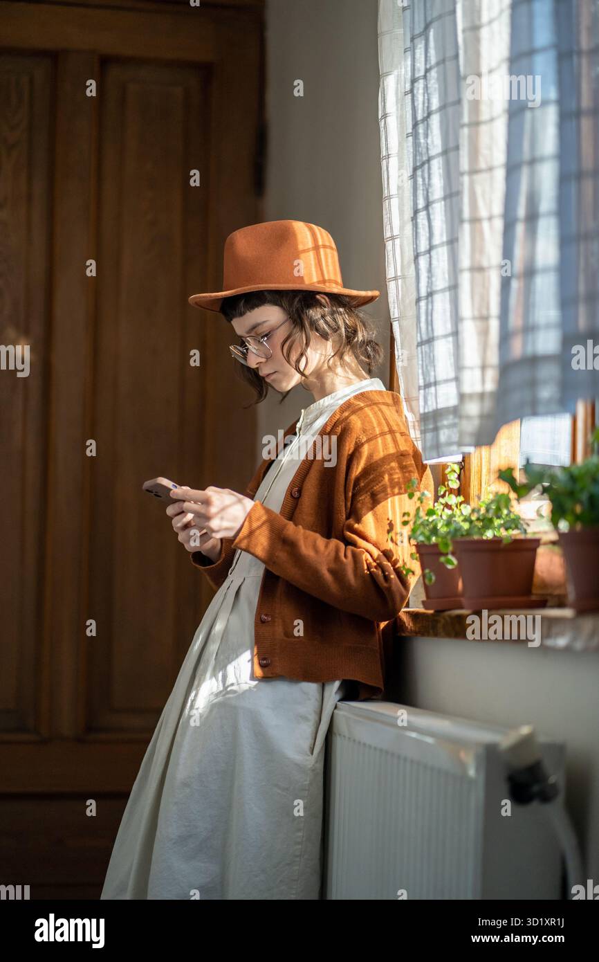 Hipster stilvolles Mädchen mit Smartphone in der Nähe des Fensters schreibt sms Verwenden Sie die mobile App, um ein Taxi nach Hause zu buchen Stockfoto