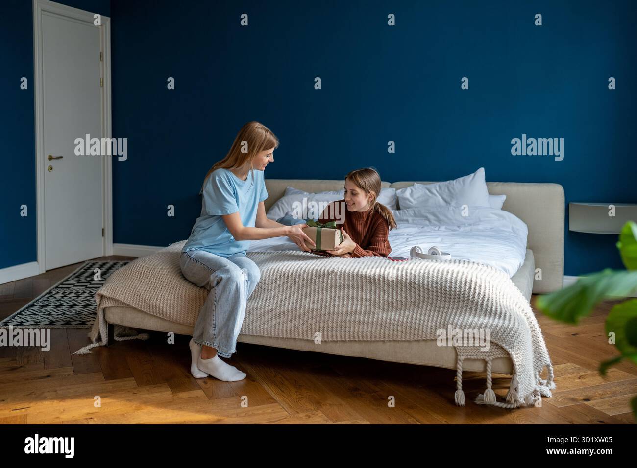 Die fürsorgliche Mutter gratuliert der fröhlichen Tochter eines Teenagers, die im Bett liegt, und schenkt eine Geschenkbox zum Geburtstag Stockfoto