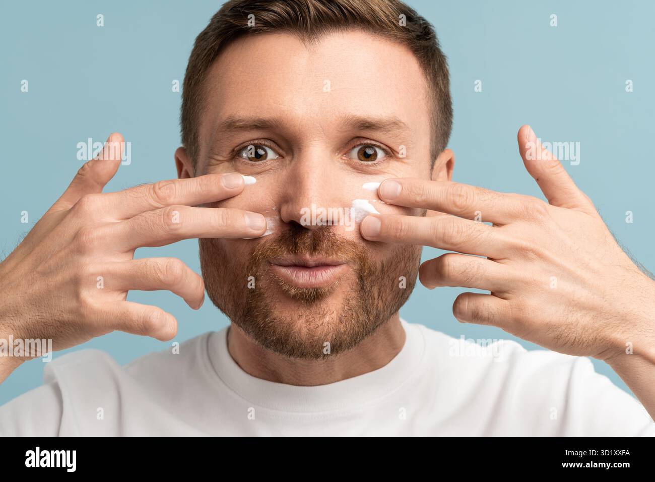 Ein junger Mann, der Spaß hat, Gesichtscreme auf die Wangen zu tragen, zwei Hände, die die Kamera anschauen, und die Nahaufnahme lächelt Stockfoto