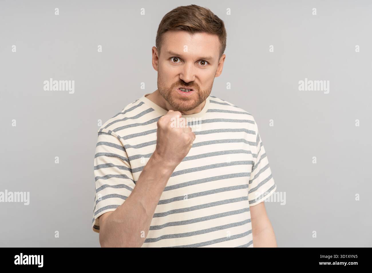 Wütender Mann zeigt Faust mit verärgerter Grimasse und verrückten Augen, die auf die Kamera schauen, isoliert an einer grauen Wand Stockfoto