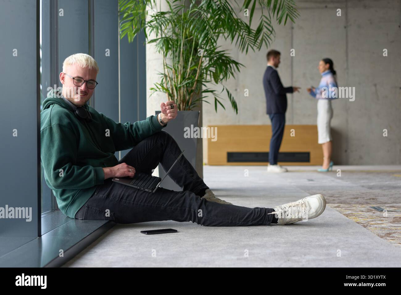Ein ungezwungener junger Profi, der in einem modernen Büro an einem großen Fenster sitzt, lächelt, während er an einem Laptop arbeitet, während Kollegen unscharf interagieren Stockfoto
