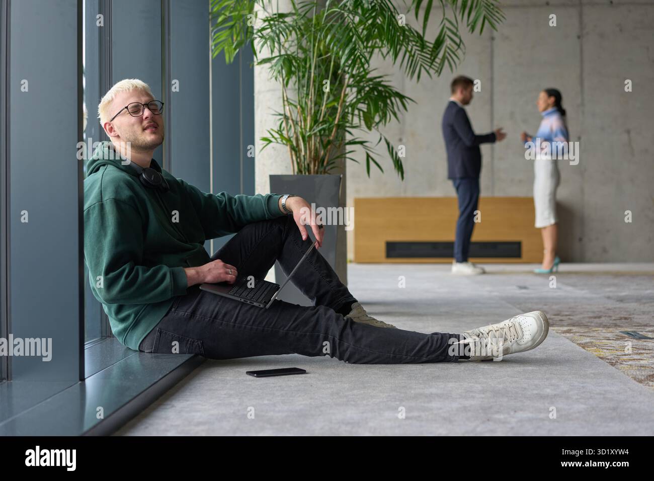 Ein ungezwungener junger Profi, der in einem modernen Büro an einem großen Fenster sitzt, lächelt, während er an einem Laptop arbeitet, während Kollegen unscharf interagieren Stockfoto