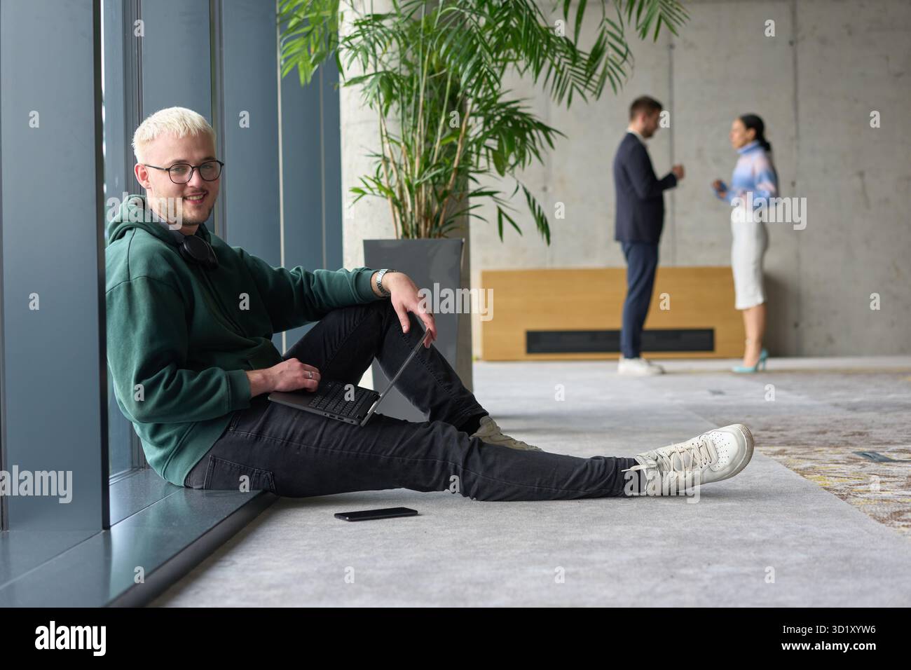 Ein ungezwungener junger Profi, der in einem modernen Büro an einem großen Fenster sitzt, lächelt, während er an einem Laptop arbeitet, während Kollegen unscharf interagieren Stockfoto