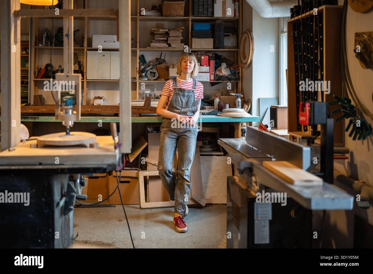 Entspannte Tischlerin, die Zeit am Arbeitsplatz verbringt, macht Pause in der Tischlerei mit Blick auf die Kamera. Stockfoto